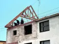 Trzech budowlańców pracuje przy podnoszeniu drewnianej konstrukcji dachu. Widać postęp prac budowlanych.