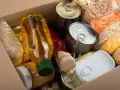 Pudełko z zapasami żywności długoterminowej: puszki, ryż, soczewica, płatki owsiane, olej i inne produkty.