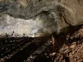 Wnętrze jaskini ciemnej, z kamienistym dnem i skalnymi ścianami. W oddali widać dwie osoby zwiedzające.