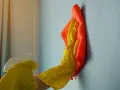 Ręka w żółtej rękawiczce z pomarańczową ściereczką czyści ścianę. Dowiedz się, czym zmyć farbę ceramiczną, by przywrócić jej blask.