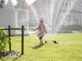 Lekfull **tipspromenad barn** i sommarregn från en vattenspridare. Ett litet barn i blommig klänning njuter av vattnet på gräsmattan.