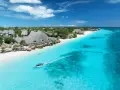Rajskie zanzibar plaże z turkusową wodą, biały piasek i łódź motorowa. W tle tropikalna roślinność i zabudowania.