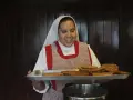 A smiling nun in a white habit and checkered apron holds a tray of fried pastries, perhaps answering the question: what do nuns eat for breakfast?