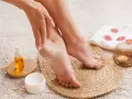 Application d'une crème hydratante sur la peau sous les pieds pour un soin doux. Une bougie parfumée et une serviette décorée complètent ce moment de détente.