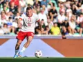 Jakub Kozłowski w akcji, w białej koszulce z orłem i czerwonych spodenkach, prowadzi piłkę na boisku.