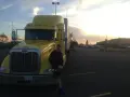 Młody mężczyzna opiera się o żółtą ciężarówkę. W tle zachód słońca i parking pełen aut. To obrazek z życia polskie truckerki.