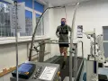 Mężczyzna w masce biegnie na bieżni podczas testów wydolnościowych. Urządzenia monitorują jego parametry życiowe.