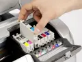Dłoń wkłada kartridż do drukarki atramentowej. Widać kolorowe tusze i mechanizm budowy drukarki.