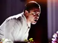 Mike Patton heute, mit weit aufgerissenem Mund und intensivem Blick, trägt ein weißes Hemd. Blumen im Vordergrund.