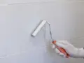 Mano enguantada pintando con rodillo blanco sobre azulejos grises. Renovando el aspecto de los tubos calefacción.