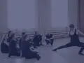 Choreograf demonstruje ruchy grupie tancerzy siedzących na podłodze.