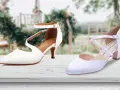Due eleganti scarpe da sposa bianche, una con cinturino alla caviglia, l'altra con incroci sul collo del piede, su un tavolo di legno con sfondo sfocato di un matrimonio.