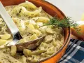 Cremiges Schmorgurken Rezept mit Dill in einer Tonschale, daneben eine Zwiebel und ein Löffel.