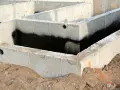 Surowe ściany fundamentowe z betonu, częściowo pokryte czarną izolacją przeciwwilgociową, otoczone ziemią.