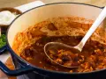 Gęste, aromatyczne chili con carne w garnku, z widoczną łyżką nabierającą porcję. Obok miseczka ze śmietaną.