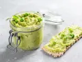 Słoik pełen zielonej, kremowej pasty z groszku, ozdobionej kilkoma groszkami i gałązką koperku. Obok kromka chrupkiego pieczywa z tą samą pastą, udekorowana kiełkami groszku.