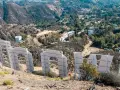 Widok zza gigantycznych liter znaku Hollywood. W oddali widać wzgórza z domami i drogami.