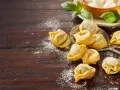Surowe tortellini posypane mąką na drewnianym blacie, obok liście bazylii i miseczka z mozzarellą.