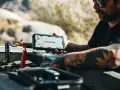 Man with tattoos demonstrates how to charge a jump starter by connecting it to a car battery in a desert landscape.