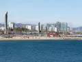 Ludzie odpoczywają na plaży, a w tle widać nowoczesne budynki Barcelony. To miasto leży nad Morzem Śródziemnym.