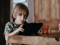 Chłopiec w hawajskiej koszuli ogląda krótkie filmy na tablecie, siedząc przy drewnianym stole.