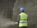Robotnik w kasku i kamizelce wykonuje torkretowanie ściany zbrojonej siatką.