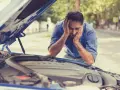 A frustrated man with hands on his head stares at his car's open hood, likely hearing a grinding noise when starting the car.
