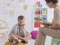 Chłopiec siedzi na poduszce, zastanawiając się, jakie pytania zadaje psycholog dziecku. W tle rysunki dzieci i kolorowe zabawki.