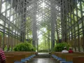 Wnętrze kaplicy w lesie, gdzie architektura organiczna łączy się z naturą. Drewniane ławki, paprocie i światło przenikające przez kratownicę tworzą spokojną atmosferę.