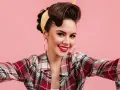 Uśmiechnięta kobieta w stylu pin-up, z burzą brązowych włosów spiętych w kok i żółtą bandaną, w kratę koszuli, robi sobie selfie.