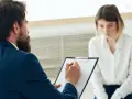 Mężczyzna z brodą w garniturze notuje coś na kartce, siedząc naprzeciwko kobiety.