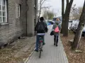 Osoba na rowerze i kobieta idąca chodnikiem obok budynku i drzew.