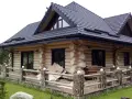 Drewniany dom z bali z czarnym dachem i tarasem, otoczony zielenią.