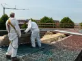 Dwóch pracowników w kombinezonach ochronnych usuwa z dachu stare płyty eternitowe. W tle widać żuraw budowlany.