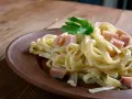 Fettuccine co to? Pyszne, kremowe fettuccine z kawałkami szynki i natką pietruszki na brązowym talerzu.