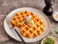 Zwei goldbraune Karotten Waffeln mit einem Klecks Sahne und geraspelten Karotten, garniert mit Kresse.