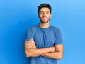 Hombre sonriente con estilos barba poco poblada, brazos cruzados, sobre fondo azul.
