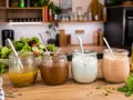 Vier Gläser mit verschiedenen salat dressing Sorten stehen auf einem Holztisch. Im Hintergrund ein bunter Salat.
