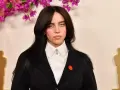 Billie Eilish z charakterystycznymi sanpaku eyes, w czarnym żakiecie i białej koszuli, na tle ozdobnych kwiatów i złotych statuetek.