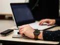 Dłoń z zegarkiem trzyma długopis nad dokumentem, obok laptop. Czas na skargę na przewlekłość postępowania opłatę.