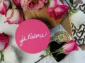 Różowe i białe róże otaczają okrągłą kartkę z napisem "je t'aime". To francuskie "dziekuje" za miłość.