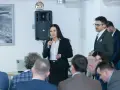 Kobieta w garniturze przemawia do grupy ludzi, obok mężczyzna w okularach.
