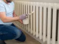 Hombre con guantes pinta radiadores blancos con brocha y pintura blanca.