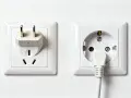 Två vita eluttag på en vit vägg. Ett med en adapter för amerikanska kontakter, det andra med en vit kabel instucken. Svenska eluttag är standard.