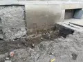 Widoczne ślady osiadania budynku i pęknięcia na cokole. Uszkodzony fragment fundamentu, ziemia i gruz wokół.