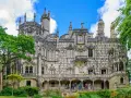 The Quinta da Regaleira estate in Sintra, Portugal, features a stunning palace with intricate stonework and ornate balconies, surrounded by lush greenery under a blue sky.