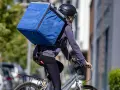 Kurier rowerowy z dużą niebieską torbą na plecach, w kasku i okularach, w drodze na rozwożenie jedzenia.
