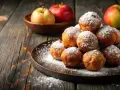 Montón de buñuelos de manzana espolvoreados con azúcar glas sobre un plato oscuro. Manzanas rojas y verdes en un cuenco de madera al fondo.