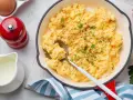 Un plat de oeufs brouillés, parsemé de ciboule et de poivre, servi dans une poêle rouge. Un moulin à poivre rouge et une cruche de lait sont à proximité.