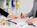 Podpisanie dokumentów, które mogą dotyczyć wypowiedzenia umowy zlecenia. Na stole leży model domu, klucze i lupa.